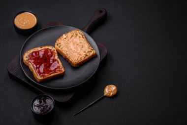 Nutritious sandwiches consisting of bread, raspberry jam and peanut butter on a black ceramic plate on a dark concrete background