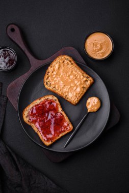 Nutritious sandwiches consisting of bread, raspberry jam and peanut butter on a black ceramic plate on a dark concrete background