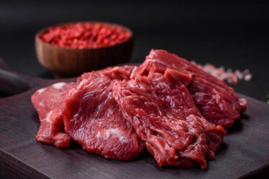Raw beef cut into several pieces on a wooden cutting board on a dark concrete background