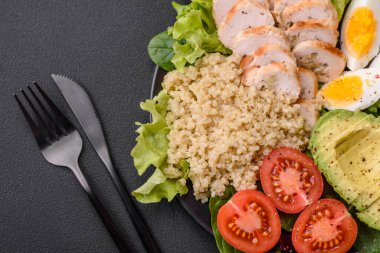 Delicious healthy lunch consisting of chicken, avocado, quinoa, eggs, tomatoes, lettuce and greens on a black plate on a concrete background