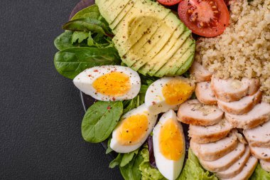 Delicious healthy lunch consisting of chicken, avocado, quinoa, eggs, tomatoes, lettuce and greens on a black plate on a concrete background