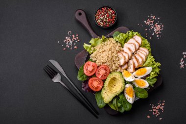 Delicious healthy lunch consisting of chicken, avocado, quinoa, eggs, tomatoes, lettuce and greens on a black plate on a concrete background
