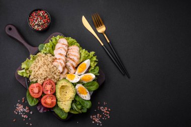 Delicious healthy lunch consisting of chicken, avocado, quinoa, eggs, tomatoes, lettuce and greens on a black plate on a concrete background