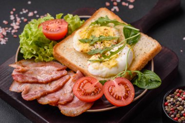 Delicious healthy lunch consisting of bacon, toast, eggs, tomatoes, lettuce and greens on a black plate on a concrete background