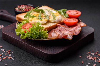 Delicious healthy lunch consisting of bacon, toast, eggs, tomatoes, lettuce and greens on a black plate on a concrete background