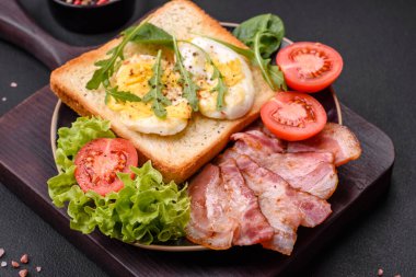 Delicious healthy lunch consisting of bacon, toast, eggs, tomatoes, lettuce and greens on a black plate on a concrete background