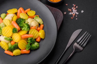 Delicious fresh vegetables steamed carrots, broccoli, cauliflower on a black plate on a dark concrete background