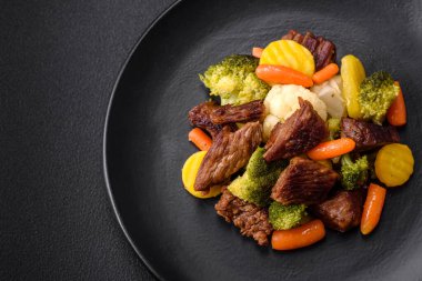 Delicious fresh beef and vegetables carrots, broccoli, cauliflower on a black plate on a dark concrete background