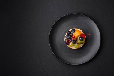 Delicious fresh tart with blueberries, cherries, grapes on a black ceramic plate on a dark concrete background