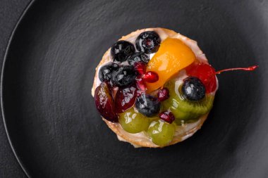 Delicious fresh tart with blueberries, cherries, grapes on a black ceramic plate on a dark concrete background