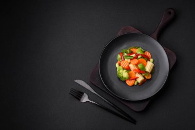 Delicious juicy broccoli vegetables, carrots, asparagus beans, bell peppers steamed in a black plate on a dark concrete background