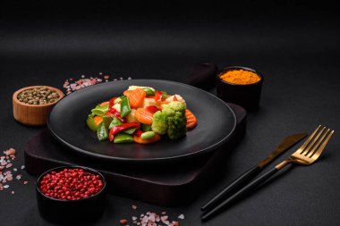 Delicious juicy broccoli vegetables, carrots, asparagus beans, bell peppers steamed in a black plate on a dark concrete background