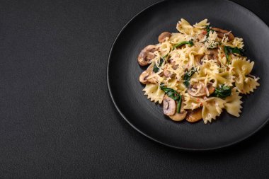 Delicious farfalle pasta with mushrooms, cheese and spinach with spices on a black plate on a dark concrete background