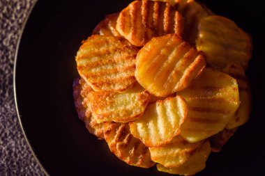 Delicious crispy fried potatoes in slices on a black ceramic plate on a dark concrete background