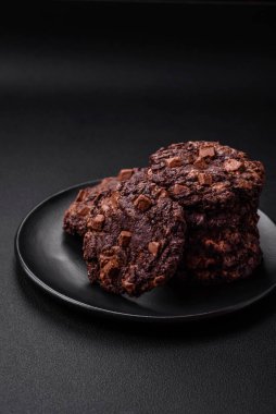 Koyu beton arka planda siyah seramik tabakta çikolatalı ve fındıklı lezzetli çıtır yulaf ezmeli kurabiyeler.