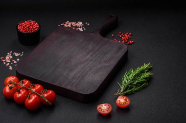 Boş ahşap kesme tahtasıyla kiraz domatesleri, baharatlar, tuz ve otlar koyu beton arka planda.