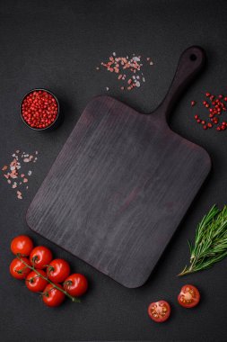 Boş ahşap kesme tahtasıyla kiraz domatesleri, baharatlar, tuz ve otlar koyu beton arka planda.