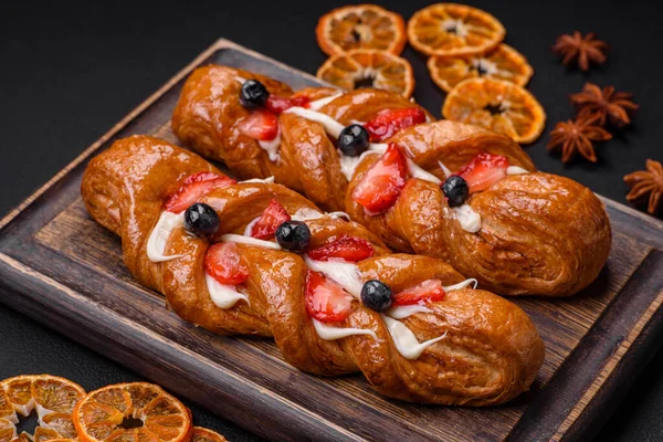 stock image Delicious fresh crispy pastry in the form of a pigtail with strawberries and blueberries on a dark concrete background