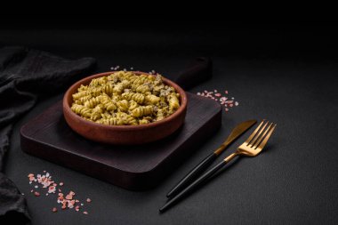 Yeşil pesto soslu lezzetli fusilli makarna, koyu beton bir zeminde seramik tabakta tuz ve baharatlar.