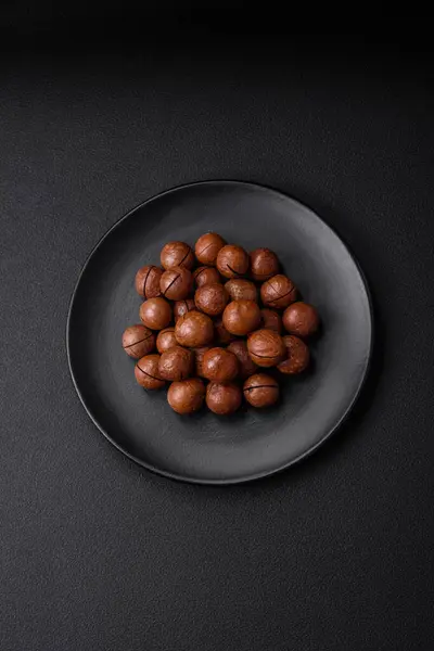 stock image Delicious roasted macadamia nuts in shell on a dark textured concrete background. Vegetarian food