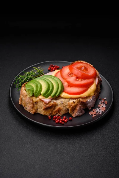 stock image Delicious nutritious grilled toast with ham, cheese, tomatoes and avocado with salt, spices and herbs on a dark concrete background