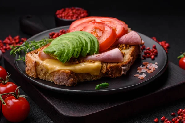 Delicious nutritious grilled toast with ham, cheese, tomatoes and avocado with salt, spices and herbs on a dark concrete background