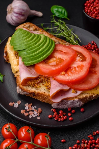 Delicious nutritious grilled toast with ham, cheese, tomatoes and avocado with salt, spices and herbs on a dark concrete background