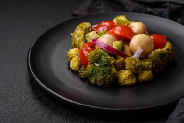 Brokolili taze salata, kiraz domatesleri, mantarlar, soğanlar, tuz, baharatlar ve koyu beton arka planda otlar.