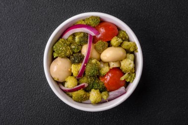 Brokolili taze salata, kiraz domatesleri, mantarlar, soğanlar, tuz, baharatlar ve koyu beton arka planda otlar.