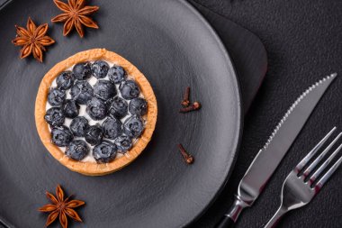 Olgun yaban mersinli ve kremalı lezzetli yuvarlak turta, koyu beton bir arka planda.