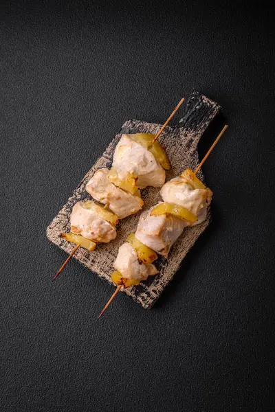 Delicious chicken or turkey kebab with pineapple pieces on skewers with salt, spices and herbs on a dark concrete background