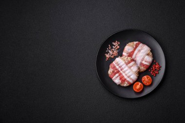 Koyu beton bir arka planda tuzlu, baharatlı ve otlu domuz pastırmasına sarılı yuvarlak doğranmış tavuk veya domuz pirzolası.
