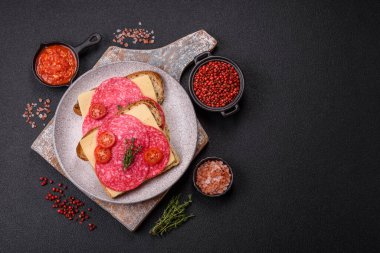 Ekmek arası sandviç, salam dilimleri, peynir ve domates, tuz ve baharatlar koyu beton arka planda.