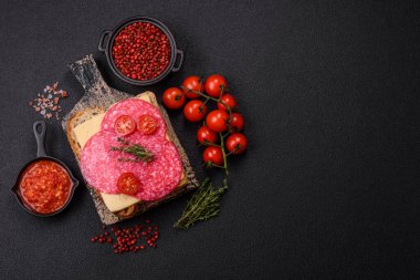 Ekmek arası sandviç, salam dilimleri, peynir ve domates, tuz ve baharatlar koyu beton arka planda.