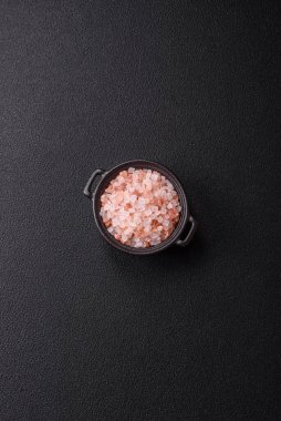 Koyu beton arka planda siyah bir kasede pembe Himalaya tuzu. Mutfakta baharatlar ve katkı maddeleri var.