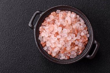 Koyu beton arka planda siyah bir kasede pembe Himalaya tuzu. Mutfakta baharatlar ve katkı maddeleri var.