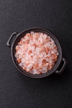 Koyu beton arka planda siyah bir kasede pembe Himalaya tuzu. Mutfakta baharatlar ve katkı maddeleri var.