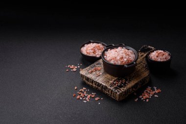 Koyu beton arka planda siyah bir kasede pembe Himalaya tuzu. Mutfakta baharatlar ve katkı maddeleri var.