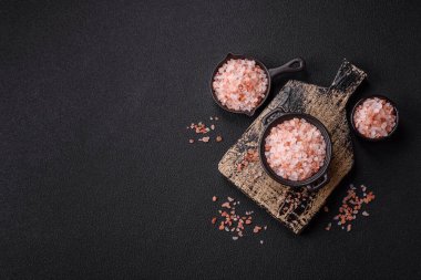 Koyu beton arka planda siyah bir kasede pembe Himalaya tuzu. Mutfakta baharatlar ve katkı maddeleri var.