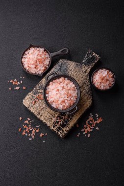 Koyu beton arka planda siyah bir kasede pembe Himalaya tuzu. Mutfakta baharatlar ve katkı maddeleri var.