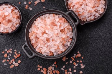 Koyu beton arka planda siyah bir kasede pembe Himalaya tuzu. Mutfakta baharatlar ve katkı maddeleri var.