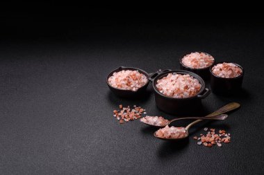 Koyu beton arka planda siyah bir kasede pembe Himalaya tuzu. Mutfakta baharatlar ve katkı maddeleri var.