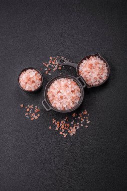 Koyu beton arka planda siyah bir kasede pembe Himalaya tuzu. Mutfakta baharatlar ve katkı maddeleri var.