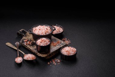 Koyu beton arka planda siyah bir kasede pembe Himalaya tuzu. Mutfakta baharatlar ve katkı maddeleri var.