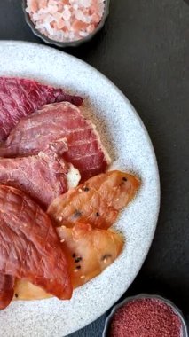 Kurutulmuş et, tuzlu, baharatlı ve otlu dilimler halinde koyu beton bir arka planda.