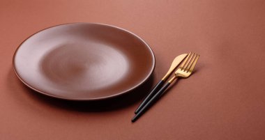 Tek renkli kahverengi arka planda boş seramik plaka. Yemek masasında çatal bıçak takımı olan boş bir tabak.