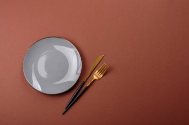 Tek renkli kahverengi arka planda boş seramik plaka. Yemek masasında çatal bıçak takımı olan boş bir tabak.