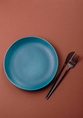 Tek renkli kahverengi arka planda boş seramik plaka. Yemek masasında çatal bıçak takımı olan boş bir tabak.