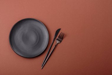 Tek renkli kahverengi arka planda boş seramik plaka. Yemek masasında çatal bıçak takımı olan boş bir tabak.