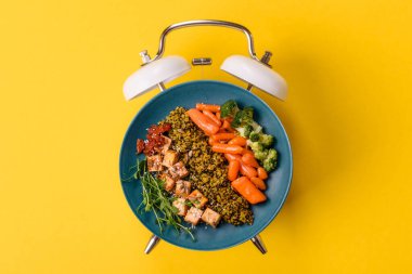 Çalar saatin arka planında kinoalı, sebzeli, etli ve yumurtalı vejetaryen salatası ya da kase. Vejetaryen yemeği yemek için kavramsal geçmiş zamanı